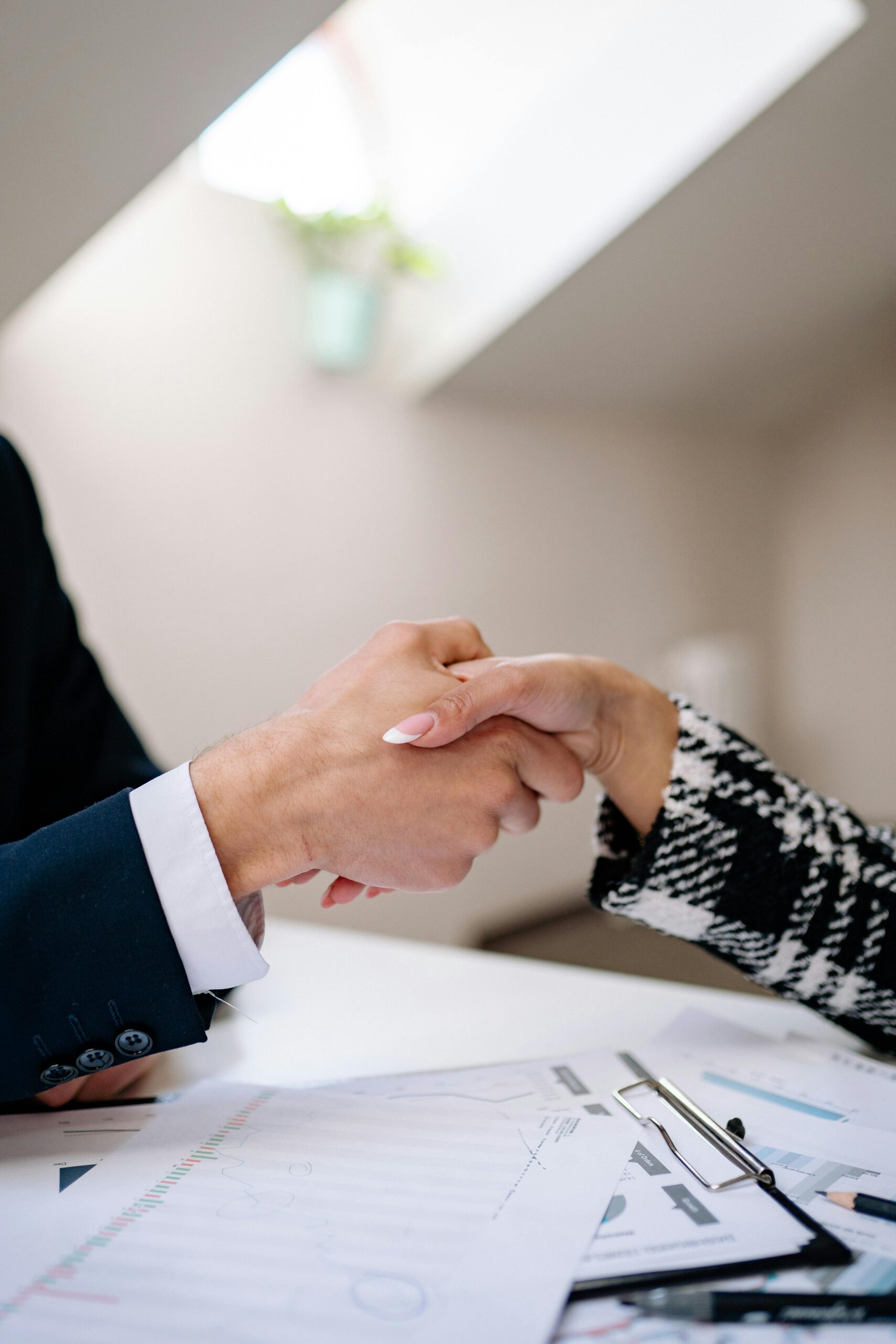 A close-up of two professionals shaking hands across a desk, with financial documents and a laptop in the background.