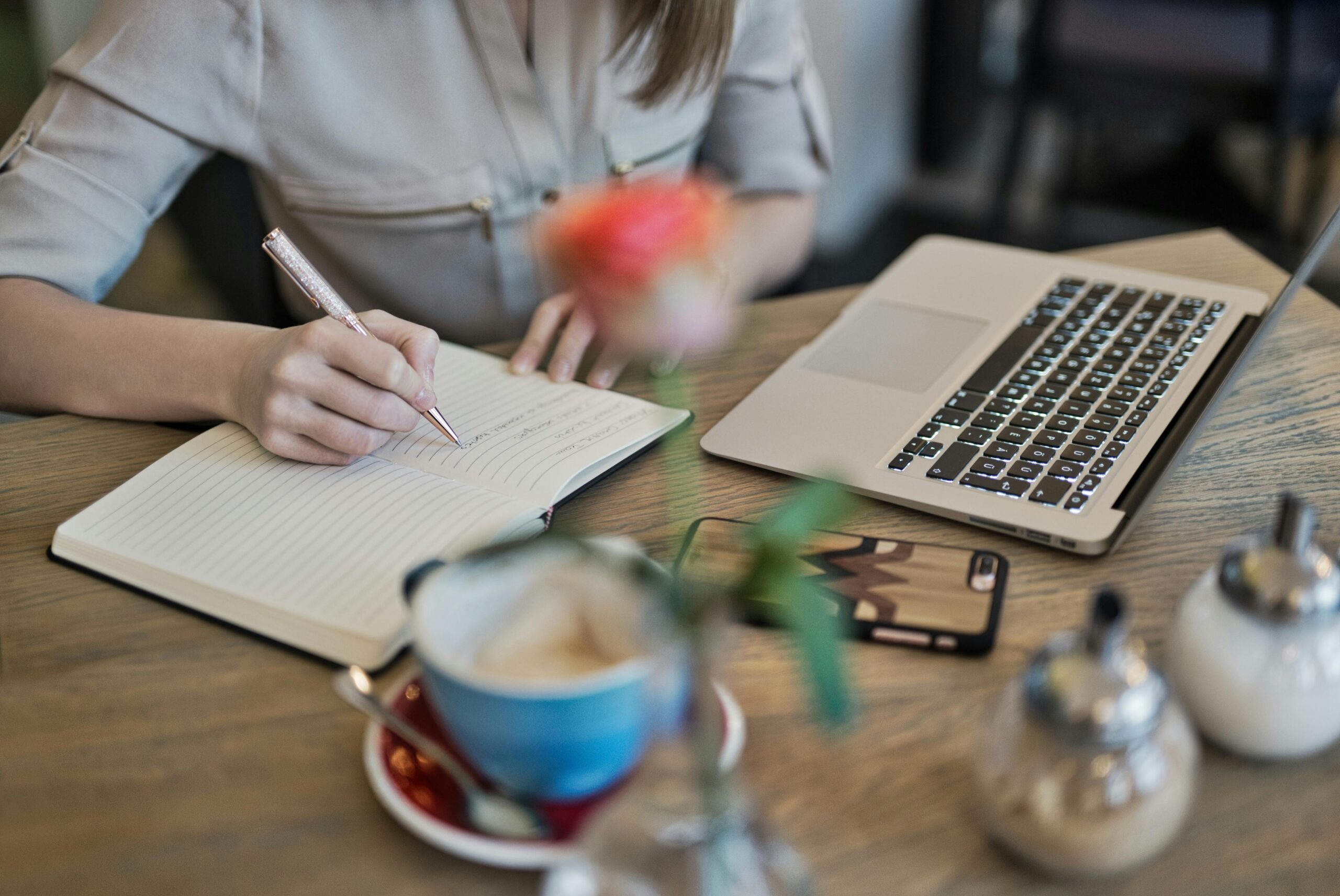 A woman writing in a notebook at a café table with a laptop, phone, and coffee.