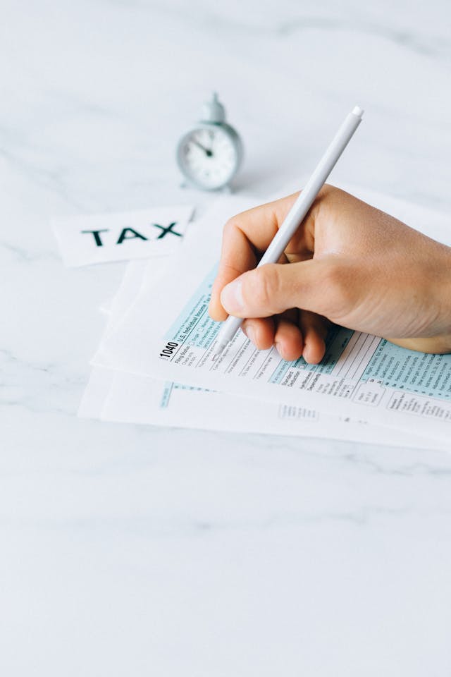 A person filling out a tax form with a pen, with a clock and "TAX" label in the background.