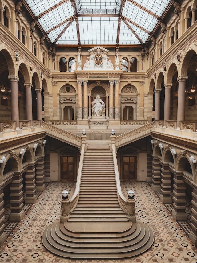 A grand, historic courthouse interior with a wide staircase leading to a statue under a glass ceiling, featuring intricate architectural details.