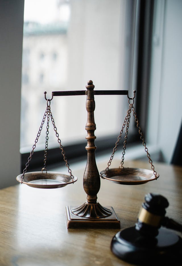 A balance scale and a gavel on a wooden table near a window.